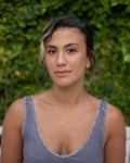 A woman with dark hair pulled back sits centered in front of a green hedge, wearing a gray ribbed tank top. Colorful outdoor pillows are visible on a white sofa behind her.