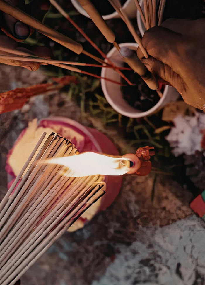 Hand lighting incense sticks with a match, surrounded by dried plants and flowers.