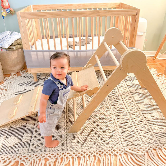 baby standing beside a wooden climber smiling and about to climb from Sapiens Child.