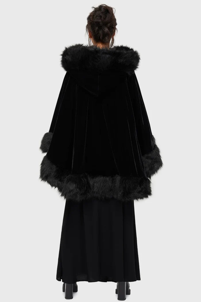 Model wearing a black faux fur-trimmed hooded cloak over a black dress on a white background