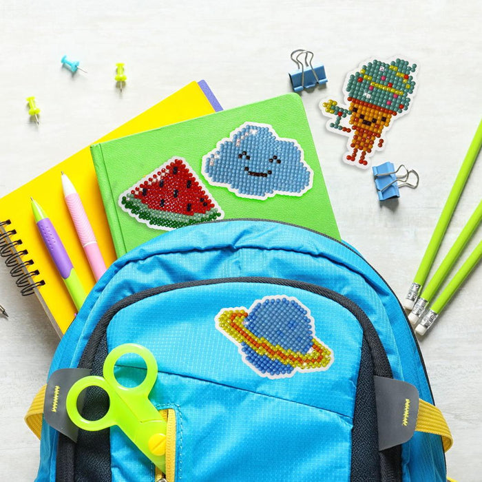 A blue backpack with colorful fuse bead patches, including a planet and Diamond Dotz Dotzies Stickers - Healthy by Diamond Dotz, sits beside green and yellow notebooks, pencils, pens, binder clips, and green scissors on a light surface.