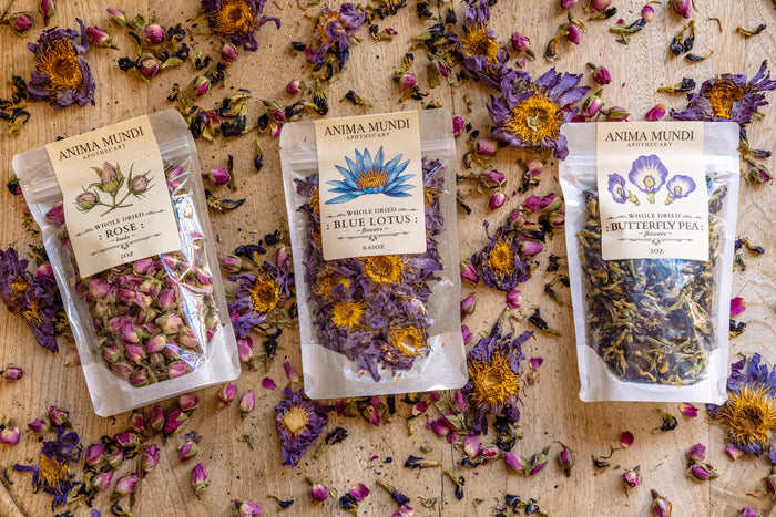 Three packages of dried flowers labeled 'Rose', 'Blue Lotus', and 'Butterfly Pea' on a wooden surface with scattered flowers.