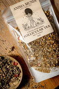 A clear stand-up pouch of Anima Mundi Apothecary Cycle Syning Follicular Tea with a beige label, shown on a rustic wooden surface with a small bowl of loose tea nearby.