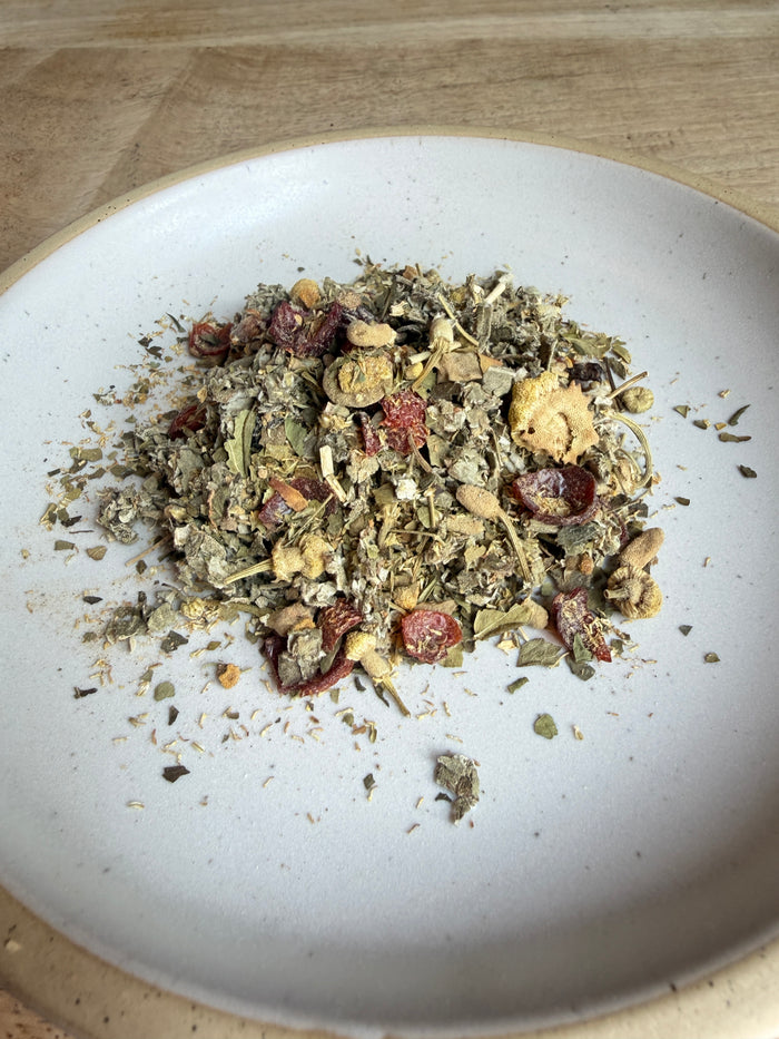 A close-up top-down view of a pile of loose herbal tea or granola blend resting in the center of a white ceramic plate, with dried green leaves, light tan pieces, and small red dried fruit pieces visible.