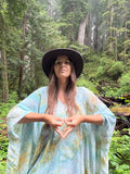 A woman wearing a wide-brimmed black hat and a light blue tie-dye kaftan poncho stands in a lush green forest, forming a heart shape with her hands.