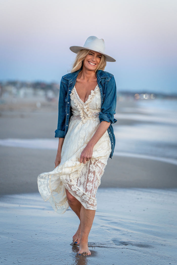 A woman walking barefoot on a beach wearing a white lace dress under a blue denim jacket and a wide-brimmed white hat.
