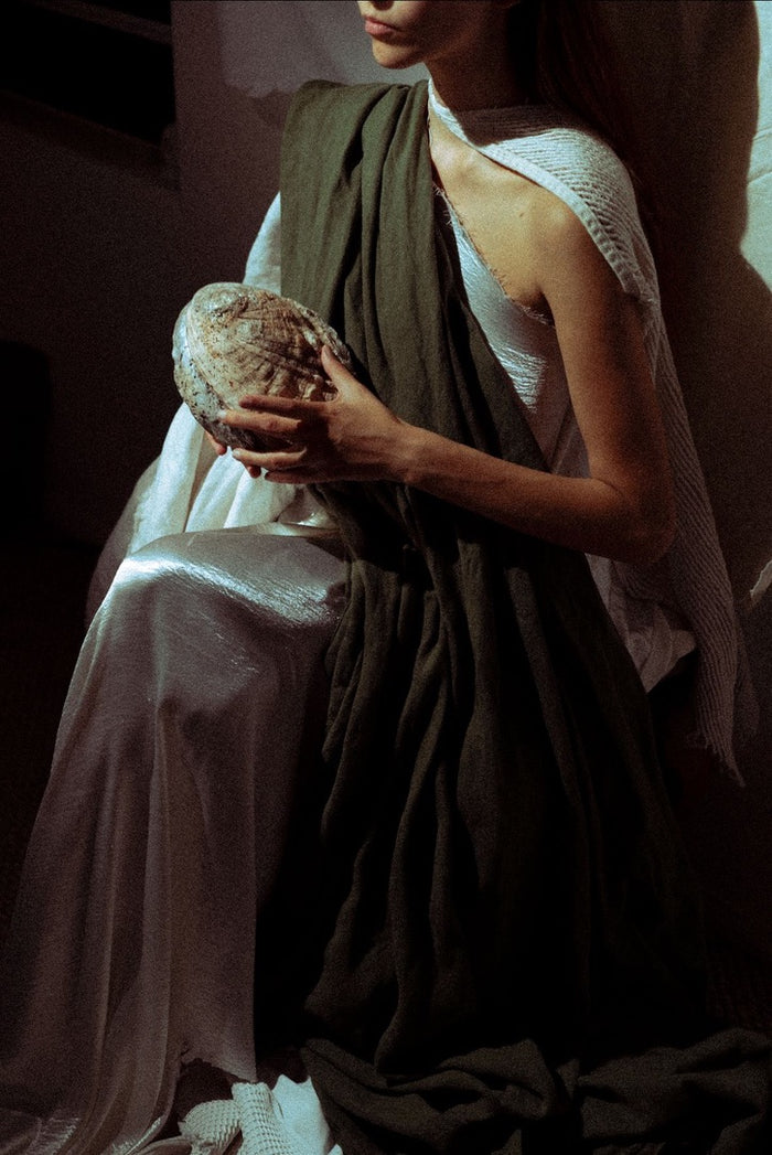 A model wearing a draped white dress with a dark olive green shawl, holding a textured shell in a dimly lit, moody setting.
