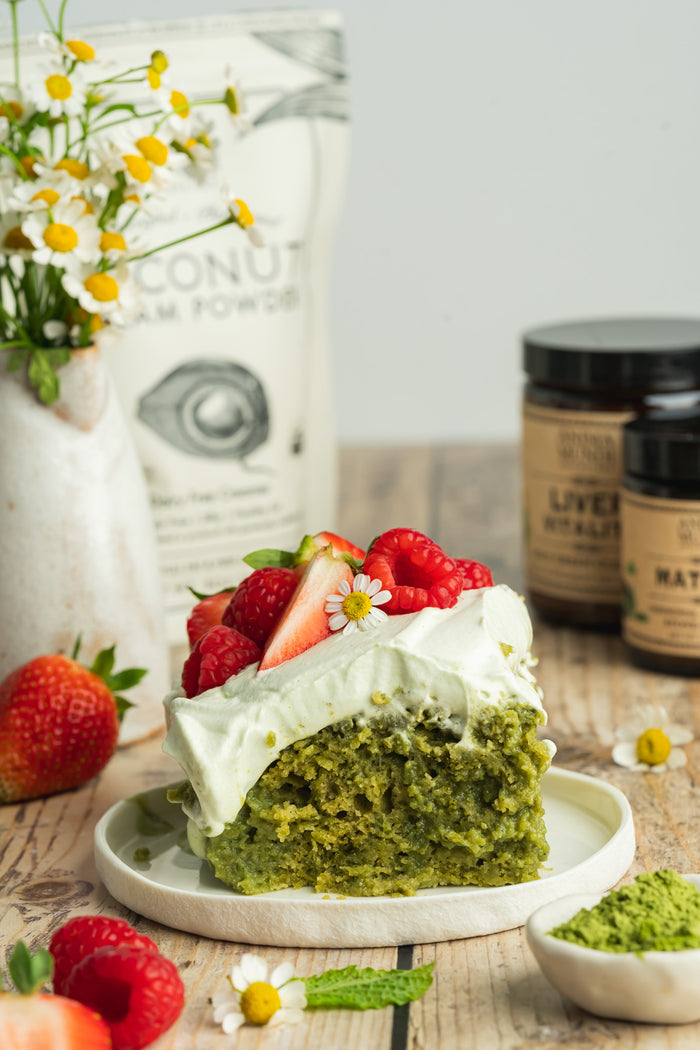 Green cake with white frosting and strawberries on a wooden table with coconut cream powder and other ingredients in the background.