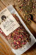 A 2oz stand-up pouch of Amundi Botanicals Cycle Syncing Moon Tea, filled with dried herbs and pink flower petals, displayed on a rustic wooden surface next to a wooden spoon filled with the tea.