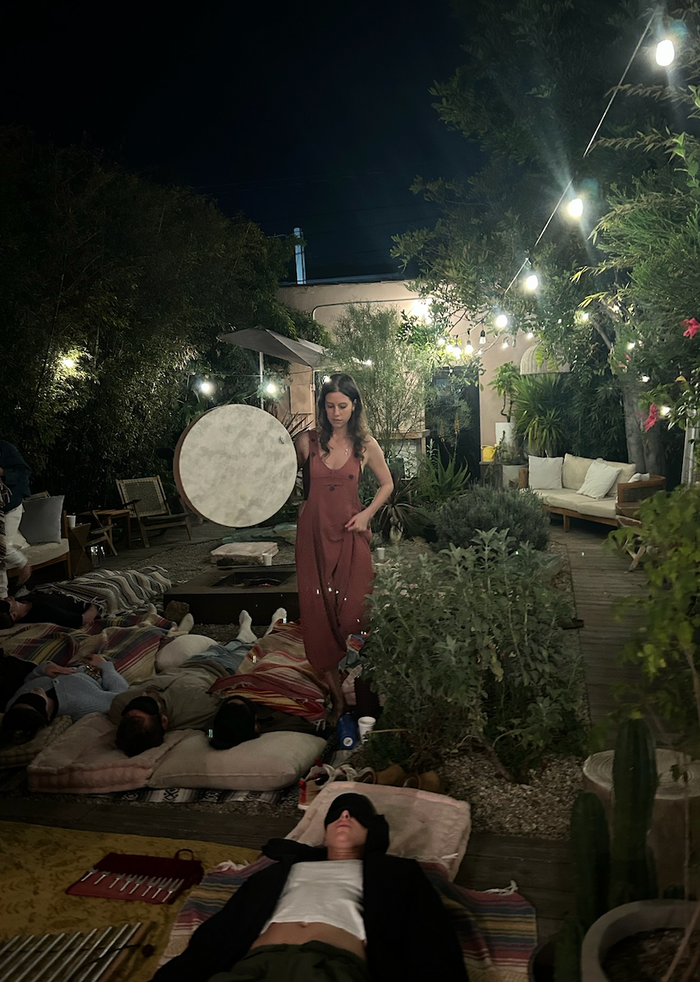 Woman in a red dress standing in a garden with people lying on the grass at night.