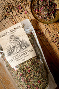 A clear plastic bag of Anima Mundi Apothecary Cycle Syncing Ovulatory Tea filled with dried herbs and pink petals, placed on a rustic wooden surface with a small dish of loose tea nearby.