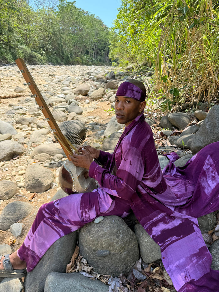 Person in purple traditional attire playing a stringed instrument by a rocky stream.
