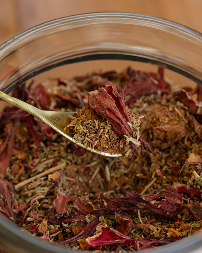 Close-up of a spoonful of dried herbs over a glass jar filled with various herbs.
