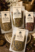 Three clear stand-up pouches of Anima Mundi botanical tea arranged on a rustic wooden surface with floral props and brass kitchenware. The visible labels read Extra Virgin Ovulatory Tea, Follicular Tea, and Luteal Tea.