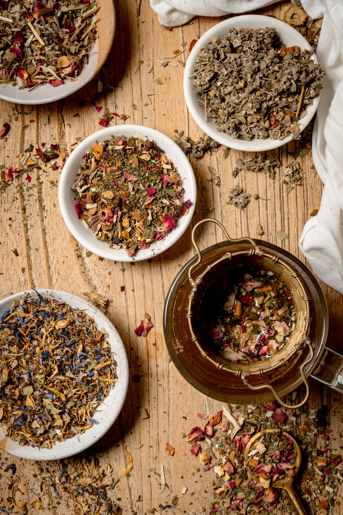 Loose-leaf herbal tea blends displayed in small white bowls and a glass teapot on a rustic wooden surface.