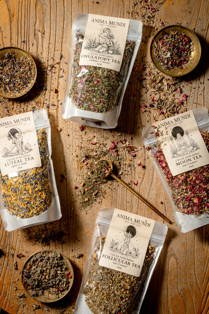 Four stand-up pouches of Anima Mundi herbal teas (Ovulatory Tea, Luteal Tea, Pollinator Tea, Moon Tea) arranged on a rustic wooden table with small bowls of loose herbal ingredients and a wooden spoon.